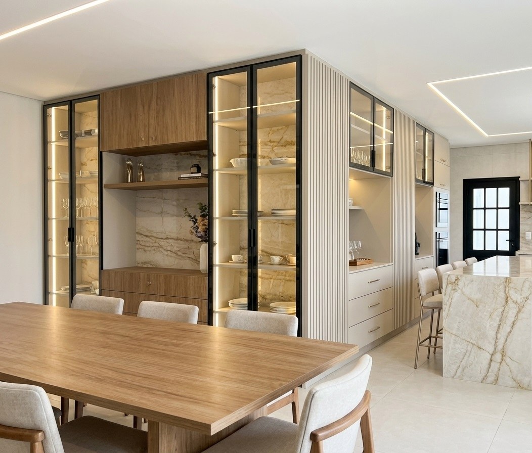 Sala de jantar com mesa de madeira diante da parede de cristaleiras iluminadas, nichos em pedra de veios dourados e painel ripado, com a ilha da cozinha ao fundo