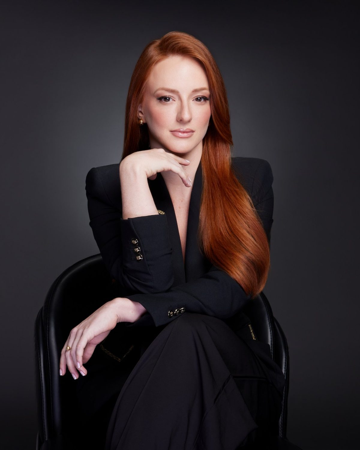 Pamela Bressan, de terno preto, sentada numa poltrona preta com a mão apoiada no queixo, sobre fundo escuro