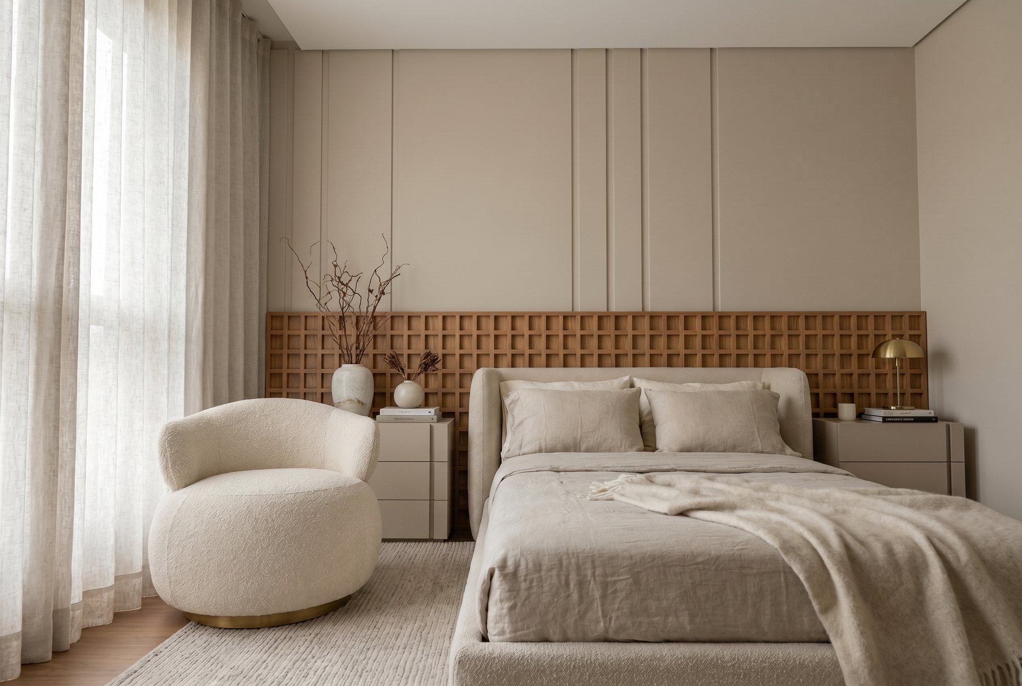 Quarto com cama estofada em bouclê, cabeceira em painel de madeira quadriculado de parede a parede, boiserie clara acima, poltrona orgânica e cortina de linho